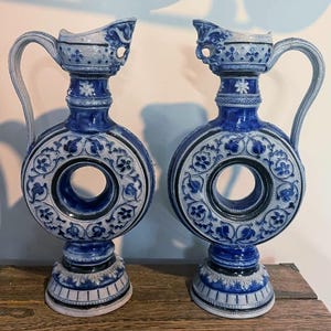 May include: A pair of matching ceramic pitchers featuring a circular, donut-shaped body with a central opening. The vessels are decorated with intricate blue and white floral relief patterns, a tall neck, a curved handle, and a wide, stable base.
