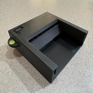 May include: A black, rectangular tool with a recessed interior and a built-in level. The tool has a series of parallel slots on one side and a small, circular level bubble. The object is made of a matte black material.