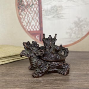 May include: Bronze-colored decorative incense burner in the shape of a turtle-dragon hybrid. The detailed sculpture features a dragon head and legs, with a turtle shell body. The burner has a lid with ornate detailing and a handle.