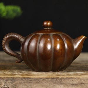 Puede incluir: Una tetera marrón con forma de calabaza, con tapa y asa curvada. La tetera tiene un pico y está hecha de un material oscuro y pulido. El asa tiene un diseño texturizado.