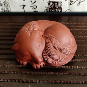 以下が含まれることがあります： テラコッタ色の丸まったキツネの彫刻。キツネは眠っている姿勢で、尾を体に巻き付けています。彫刻には、詳細な毛皮と顔の特徴があります。