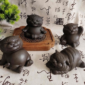 May include: Four dark brown ceramic tiger figurines in various poses. One is seated in a meditative pose on a wooden tray, while the others are in playful positions. The figurines have smiling faces and are set against a patterned background.