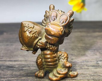 May include: A bronze-colored figurine of a cartoon dragon holding a peach. The dragon has large, expressive eyes and is seated on a wooden surface. The peach has Chinese characters on it. A yellow flower is in the background.