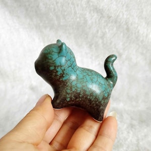 Peut inclure: Petite figurine de chat en céramique, turquoise avec un motif craquelé et des accents marron foncé. La queue est recourbée vers le haut. La figurine est tenue dans une main. Objet décoratif.