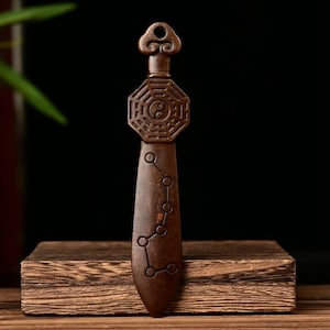 May include: A brown, intricately carved pendant with a key-like top and a geometric design featuring a yin-yang symbol. The pendant has a long, pointed body with a constellation pattern. The item is displayed on a wooden block.
