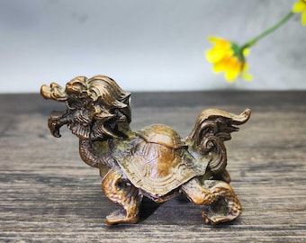 May include: A bronze-colored figurine depicting a mythical creature with a dragon-like head and a turtle shell. The sculpture has intricate details and stands on four legs. The background includes a wooden surface and yellow flowers.