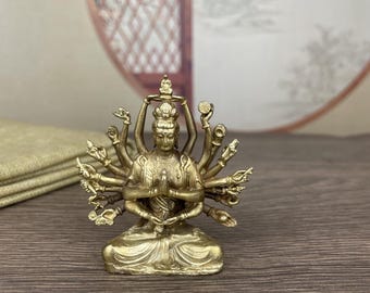 May include: A small, gold-colored statue of a seated deity with multiple arms. The figure is in a meditative pose, with hands clasped in prayer. The statue has intricate details and is likely made of metal. The background is blurred, with a hint of a decorative element.