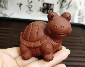 May include: A small, brown ceramic turtle figurine with a detailed shell and a smiling face. The turtle has a textured surface and is designed with a whimsical, cartoonish style. The figurine is a decorative item.