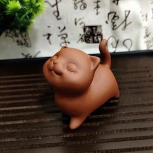 Peut inclure: Petite figurine de chat en céramique marron, les yeux fermés et une expression souriante. Le chat a une queue courbée et se tient sur une surface marron foncé. La figurine est probablement un objet décoratif.