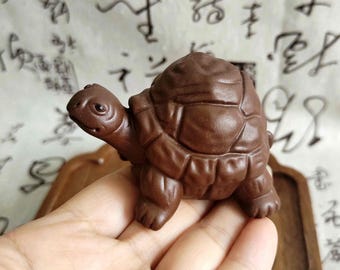 May include: A detailed, brown turtle figurine. The sculpture has a textured shell and detailed features, including a head, legs, and tail. The figurine is made of a dark brown material and is displayed on a wooden surface.