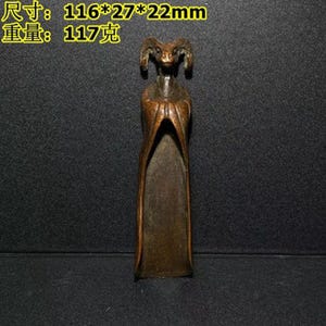 Puede incluir: Una escultura de bronce de una figura con cabeza de carnero vestida con una larga túnica. La escultura mide aproximadamente 11,6 x 2,7 x 2,2 cm y pesa 117 gramos. La figura tiene una cara y cuernos detallados.