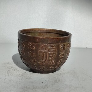 May include: A small, bronze-colored bowl with an embossed design of Chinese characters. The bowl has a rounded shape with a slightly flared rim. The characters are evenly spaced around the exterior.