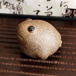 Puede incluir: Una figura de rana de cerámica marrón moteada con un solo ojo negro grande. La rana tiene un cuerpo redondeado y un pequeño bulto en la parte superior de la cabeza. La figura está sobre una superficie de madera marrón oscuro.