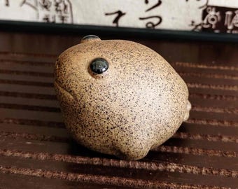 May include: A speckled, brown ceramic frog figurine with a single, large, black eye. The frog has a rounded body and a small, raised bump on top of its head. The figurine is set against a dark brown, wooden surface.
