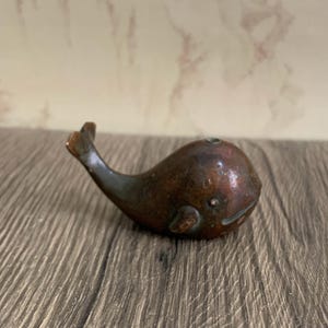 May include: A small, bronze-coloured whale figurine. The whale has a rounded body, a small tail, and a smiling face. The figurine is made of metal and has a weathered appearance. The whale is resting on a wooden surface.