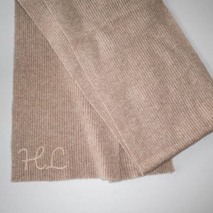 以下が含まれることがあります： ベージュのリブ編みスカーフ。明るい色合いで「HL」のイニシャルが刺繍されています。スカーフは折りたたまれており、その質感と色を示しています。ニュートラルな色合いで、汎用性の高いアクセサリーです。