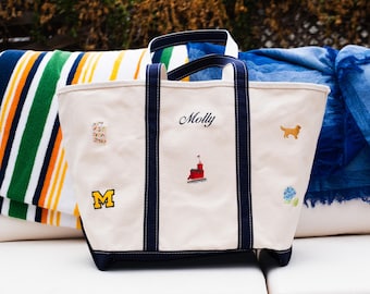 May include: A cream-colored canvas tote bag with navy blue handles and trim. The bag features embroidered designs including the name Molly, a red lighthouse, a yellow dog, a blue flower, a yellow letter M, and a small colorful patterned rectangle.
