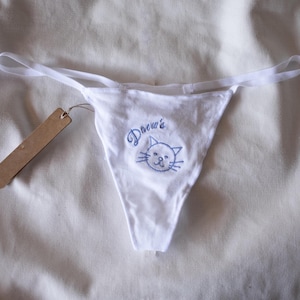 Op de afbeelding: Witte string-slip met een blauw geborduurd kattengezicht en de naam "Drew's". Het ondergoed heeft dunne witte bandjes en een klein bruin label.