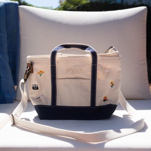 Può includere: Borsa tote color crema con dettagli blu navy e tracolla bianca. La borsa presenta ricami di una fotocamera, fiori, un gabbiano, una conchiglia e un tramonto. Le iniziali "PDE" sono ricamate.