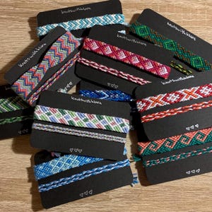 May include: A collection of handmade friendship bracelets in various colors and patterns, displayed on black cards. The bracelets feature intricate woven designs in shades of blue, pink, green, and orange. Each card has the text "knots with klara" and three heart symbols.