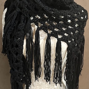 May include: A black crocheted shawl featuring an openwork pattern and long, dangling fringe along the edges. The textile accessory is draped over a white garment, showcasing the intricate yarn loops and the uniform dark color of the material.