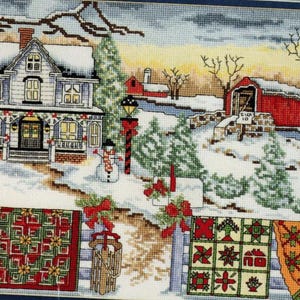 May include: A cross-stitch pattern featuring a winter scene with a house, a covered bridge, and a snowman. The house is white with a red roof and a porch. The bridge is red with a white roof. The snowman is white with a black hat and a carrot nose. The scene is surrounded by trees and snow.
