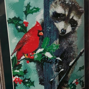 May include: A red cardinal perched on a branch with green holly and red berries, next to a raccoon peeking out from behind a tree trunk. The background is a light blue and green.