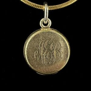 Puede incluir: Un collar de cadena de oro con un colgante redondo en forma de medallón. El medallón está grabado con un monograma.