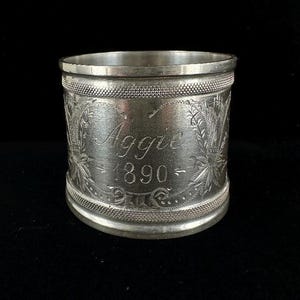 Puede incluir: Anillo de servilleta de plata con un diseño grabado y el texto "Aggie 1890".