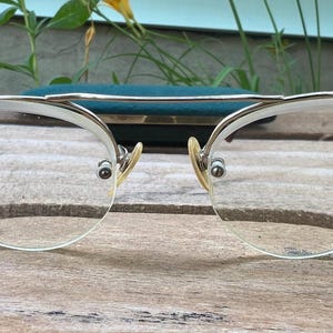 Può includere: Un paio di occhiali con montatura in metallo color argento, una barra superiore pronunciata e lenti trasparenti. Il design presenta uno stile semi-cerchiato in cui le lenti sono fissate con piccole viti sui bordi esterni e sul ponte, con naselli beige.