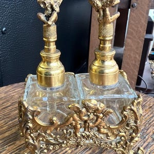 Puede incluir: Un juego de frascos de perfume vintage con dos frascos de vidrio transparente. Los frascos tienen tapones dorados ornamentados con figuras de querubines. Los frascos se asientan en una base de filigrana dorada con detalles de querubines y volutas.