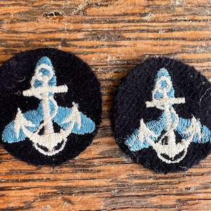 Op de afbeelding: Twee ronde zwarte stoffen patches met een geborduurd ankerontwerp. Het anker is gestikt met lichtblauw en wit garen, gecentreerd op elke ronde patch. Het borduurwerk toont een gedetailleerde ankervorm met een touw eromheen gewikkeld.