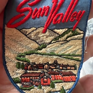 May include: A shield-shaped embroidered patch featuring a light blue sky with the red cursive text Sun Valley above a landscape of tan mountains, green trees, and a cluster of red buildings with dark roofs, all bordered by a dark blue stitched edge.