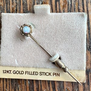 Op de afbeelding: Een goudgevulde reversspeld met een ronde, opalescente steen in een decoratieve zetting. De speld wordt gepresenteerd op een crèmekleurige stof, met de tekst "12kt. Gold Filled Stick Pin".