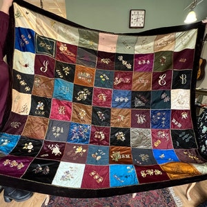 May include: A vintage patchwork quilt with embroidered letters and floral motifs. The quilt is made from a variety of fabrics in shades of brown, red, blue and green. The quilt is edged with a black velvet trim.