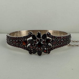 Op de afbeelding: Zilverkleurige scharnierende armband met een centraal bloemmotief. De armband is versierd met talrijke donkerrode edelstenen, met een cluster van grotere stenen in het midden. De band is afgezet met kleinere edelstenen.