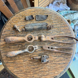May include: A collection of antique rusty metal tools and leather pieces on a wooden surface. The tools include a pair of pliers, a small wrench, and a pair of scissors. The leather pieces are brown and have a worn appearance.