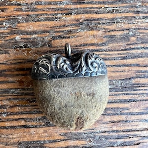 May include: A pendant shaped like an acorn featuring a textured, light brown natural nut base and a dark, oxidized silver cap with intricate, embossed swirling patterns and a small metal loop at the top for attachment to a chain or cord.