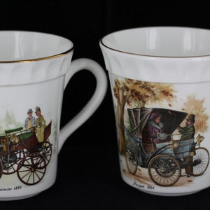 Può includere: Due tazze in ceramica bianca con bordi e manici dorati. Ogni tazza presenta un'illustrazione di auto d'epoca. Una mostra una Daimler 1896, l'altra una Peugeot 1894. Le tazze hanno un design scanalato vicino al bordo.