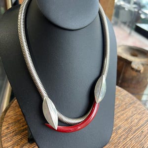 Puede incluir: Un collar de cadena de serpiente plateada con dos detalles pulidos en forma de hoja plateada y un elemento curvo y liso en forma de media luna roja, posicionado en el centro de la cadena, creando un aspecto estructurado y superpuesto.