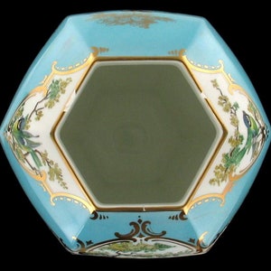 May include: A hexagonal porcelain bowl with a light blue glaze and gold trim. The bowl features a floral design with birds and green leaves. The inside of the bowl is white.