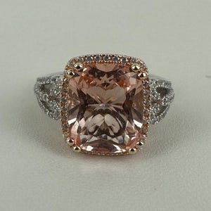 May include: A stunning engagement ring featuring a large, rectangular peach-colored gemstone. The gemstone is surrounded by a halo of smaller, clear stones set in a gold and silver band. The ring's design is elegant and eye-catching.