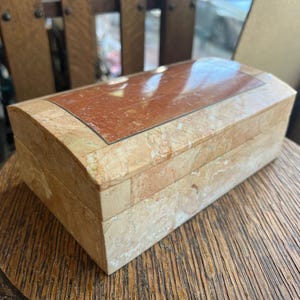 May include: A rectangular jewellery box with a curved top. The box is made of beige stone with a reddish-brown stone inlay on the lid. The box is on a wooden table.