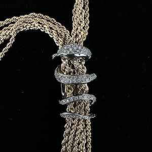 May include: A gold-coloured necklace with multiple strands, featuring a silver-coloured snake design adorned with small, sparkling stones. The snake wraps around the necklace, creating a unique and elegant look. The background is black.