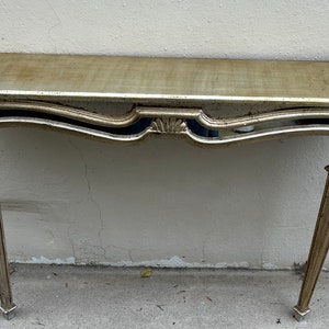 May include: A gold-toned console table with a mirrored shelf and ornate detailing. The table has a rectangular top and four slender, tapered legs. The design features curved lines and decorative carvings, creating an elegant, vintage aesthetic.