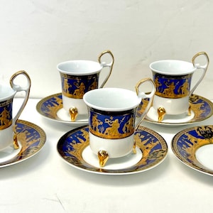 Puede incluir: Juego de cuatro tazas de espresso y platillos dorados y azules con un diseño de escena mitológica griega. Las tazas tienen tres patas doradas.