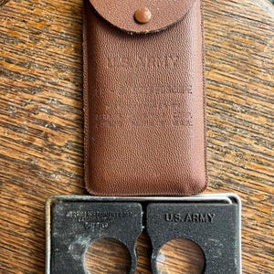 May include: A vintage US Army magnifying glass with two lenses in a brown leather case. The case has a flap with a button closure and the text "U.S. ARMY" embossed on the front. The magnifying glass has the text "ADJUSTING INSTRUMENT GOOD LENS USED" and "DATE INSPECTED" on the side.