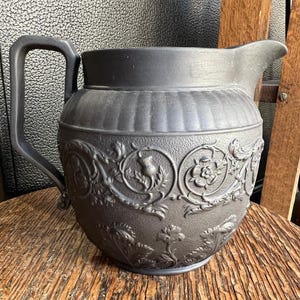 May include: A dark gray pitcher with a handle and spout, featuring embossed floral and thistle designs. The pitcher sits on a wooden surface. The pitcher's height is approximately 8 inches.