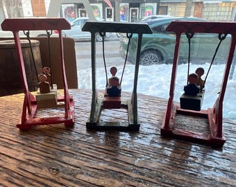 Op de afbeelding: Drie kleine houten schommels met figuren. De schommels zijn rood, groen en rood geschilderd. Elke schommel heeft een klein houten figuur dat op de schommel zit. De schommels staan op een houten tafel.