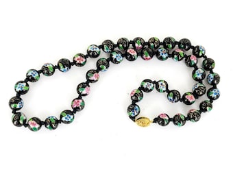 May include: A black beaded necklace with floral designs in pink, blue, and green. The beads are strung on a black cord and have a gold clasp.
